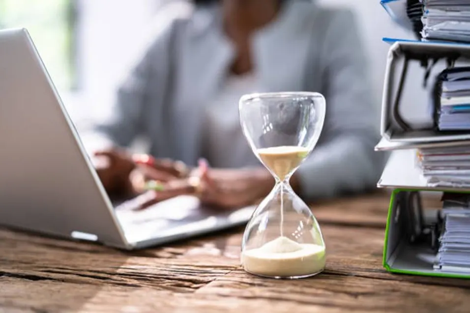 Hourglass on a wooden desk beside a laptop and a tall stack of binders, with a person working in the background
