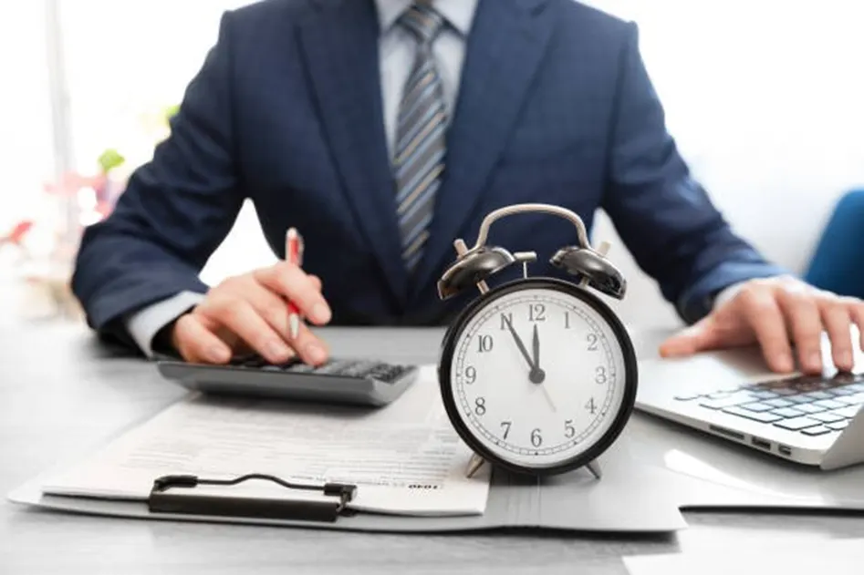 Man in a navy suit using a calculator and laptop at a desk with a large alarm clock and clipboard in the foreground