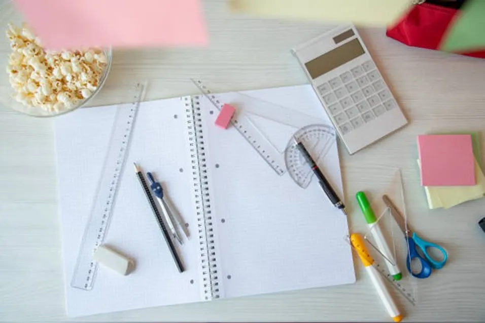 Overhead view of a gridded notebook with rulers, compass, protractor, eraser, calculator and coloured markers on a white desk
