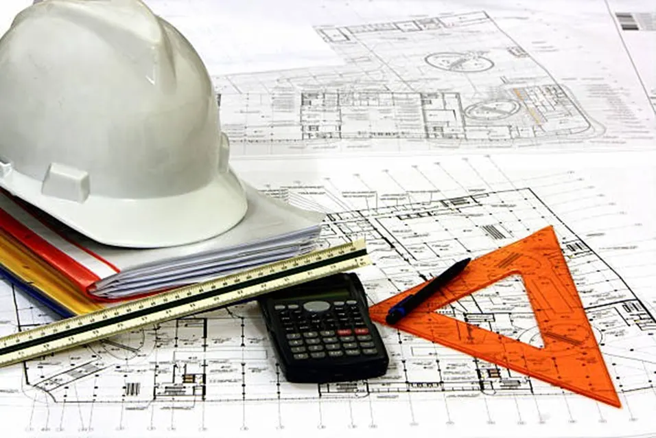 White hard hat, ruler, scientific calculator and orange set square on top of engineering blueprints