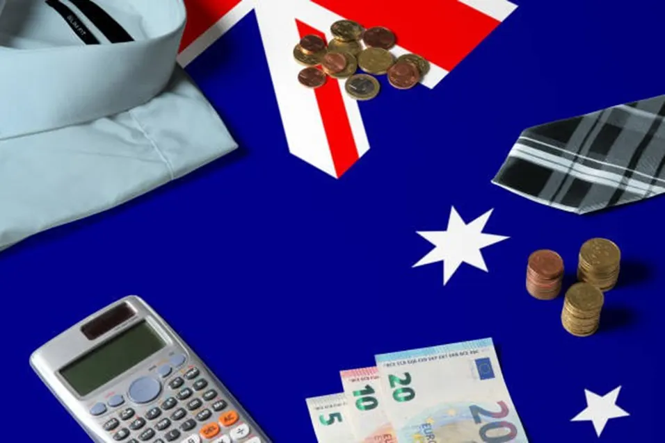 Australian flag laid flat with a calculator, euro banknotes, coins, a shirt, and a tie arranged around it