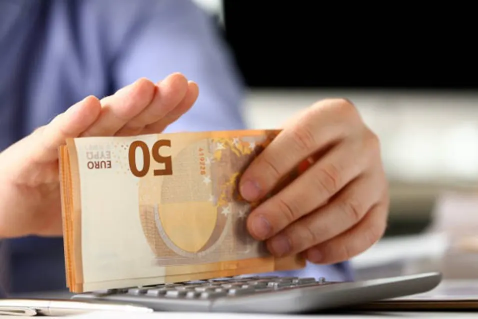 Person counting a stack of 50 euro banknotes over a calculator at an office desk