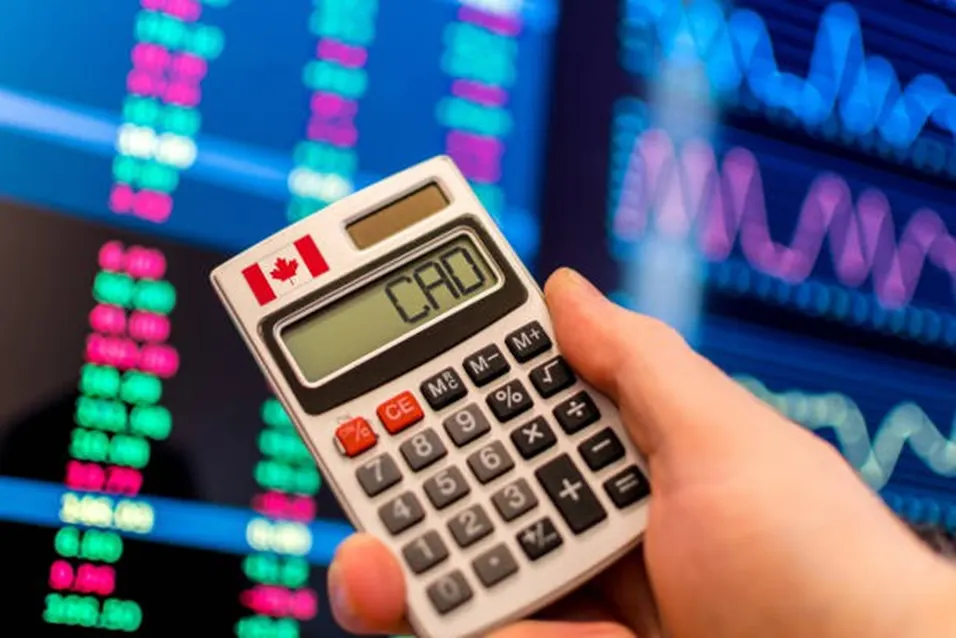 Hand holding a small calculator with the Canadian flag and CAD on its display against a stock market screen