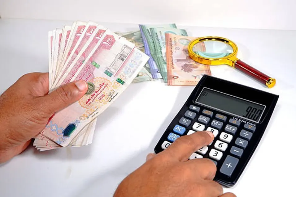Person holding a fan of UAE dirham banknotes while pressing a calculator with a magnifying glass nearby