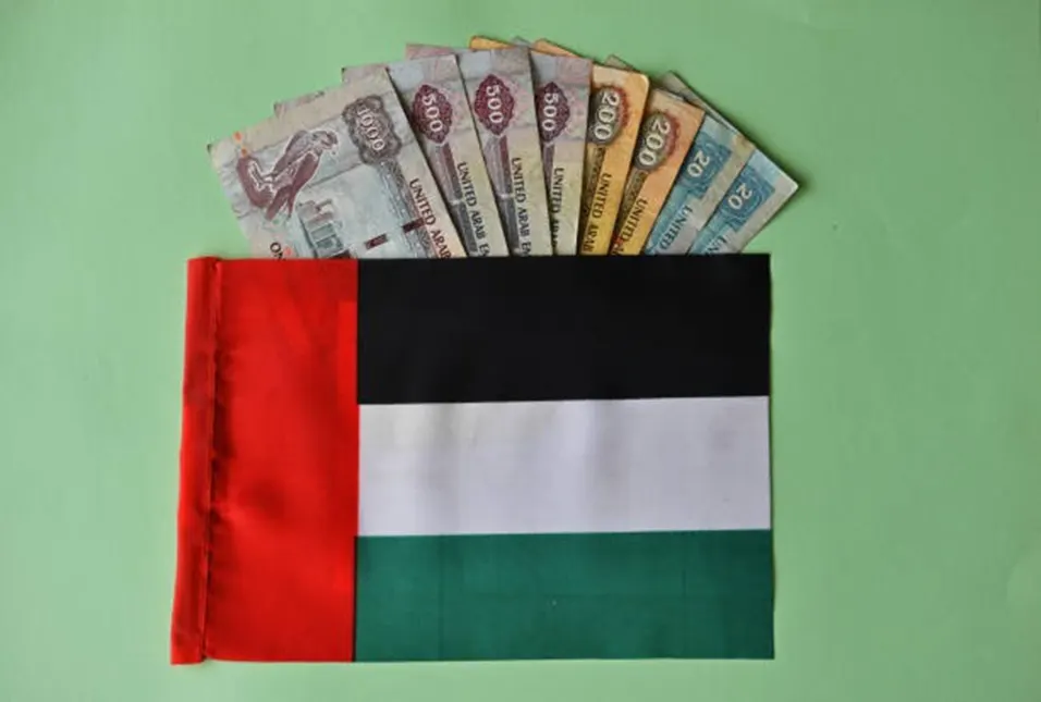 UAE flag with a spread of United Arab Emirates dirham banknotes fanned out above it on a green background