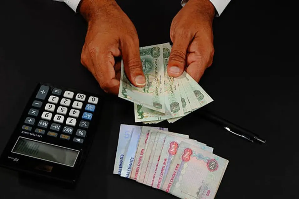Hands counting UAE dirham banknotes beside a calculator and pen on a dark desk