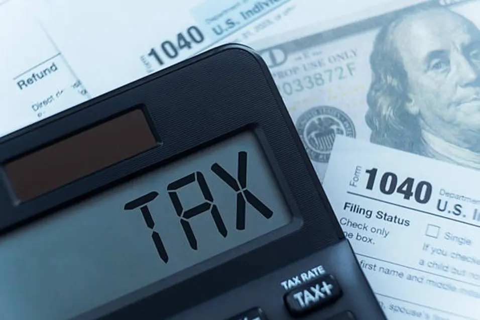 Calculator displaying TAX on its screen next to a US 1040 form and a hundred dollar bill