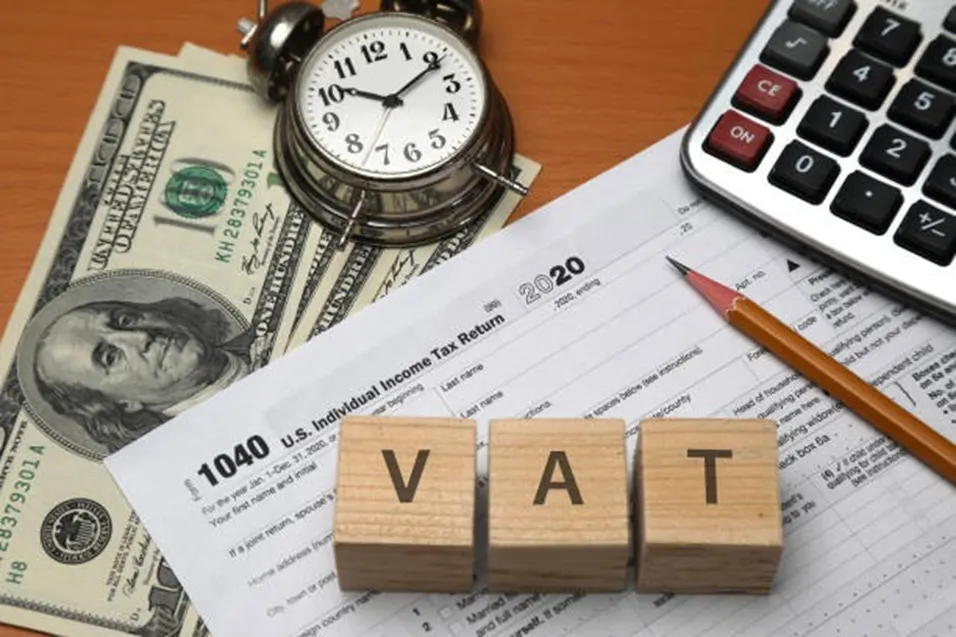 VAT wooden letter blocks beside US 1040 income tax return form, dollar bills, a clock, pencil, and calculator